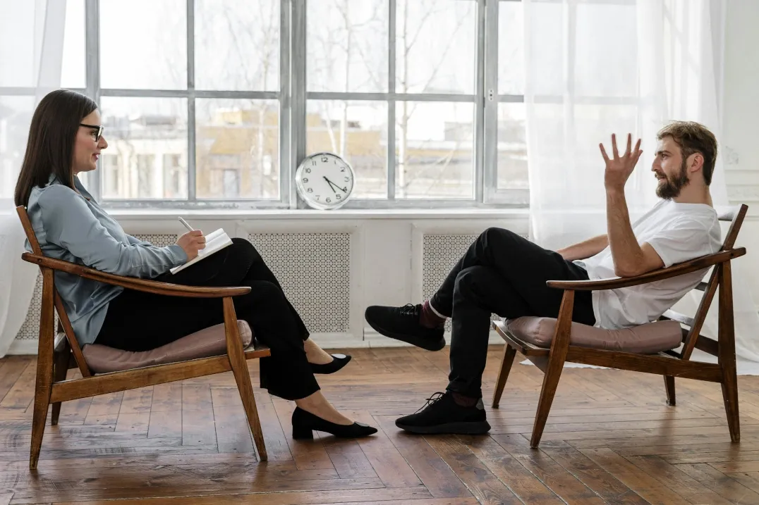 Female coach attentively listening and taking notes during a one-on-one coaching session with a male client, highlighting a focused and supportive coaching environment.