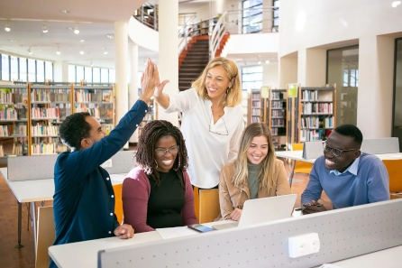 Diverse group in a coaching session celebrating success with a high-five, featuring a mentor guiding students in a library setting, highlighting teamwork and collaborative learning.