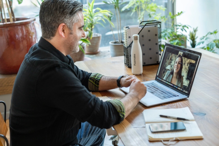 Male coach conducting an online coaching session with a client on a video call, sitting at a desk with a laptop, notebook, and plants in the background, highlighting remote coaching and virtual mentorship.