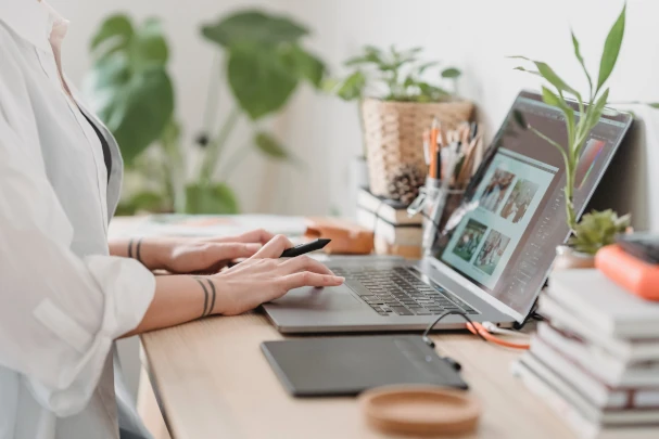 Close-up of a creative professional working on a laptop at a home office, surrounded by plants and design tools, symbolizing the focus on building a personal brand and attracting more clients.