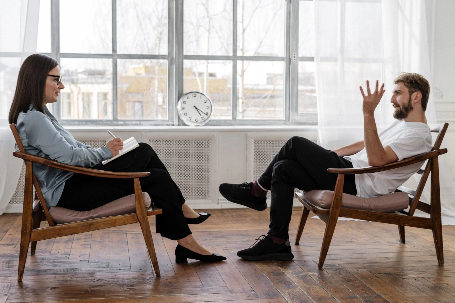 Professional coaching session with a smiling female coach taking notes while talking to a relaxed male client in a bright, airy room with large windows.