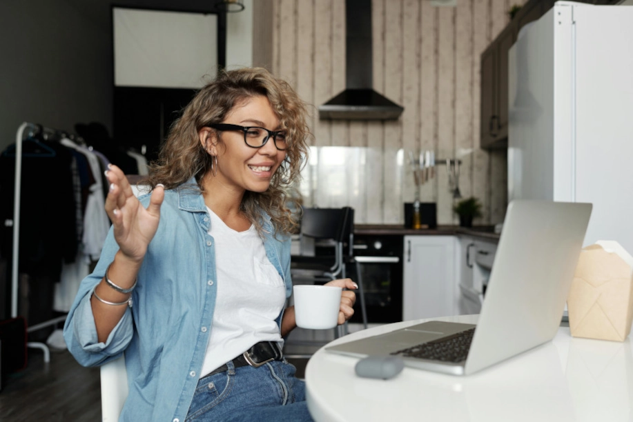 Female coach conducting an online coaching session from home, engaging with a client on a laptop while holding a coffee cup, representing remote coaching and virtual mentorship.