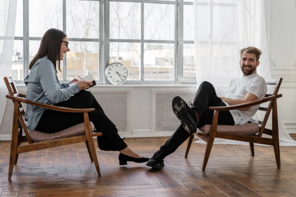 Female coach engaging in a relaxed and friendly coaching session with a smiling male client in a bright, modern room, highlighting a comfortable and supportive coaching environment.