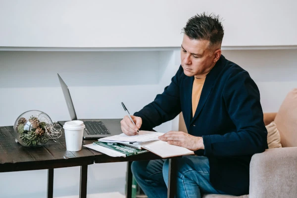 Male coach reviewing notes during a focused work session, seated at a desk with a laptop, notebook, and coffee, highlighting dedication and preparation in coaching.